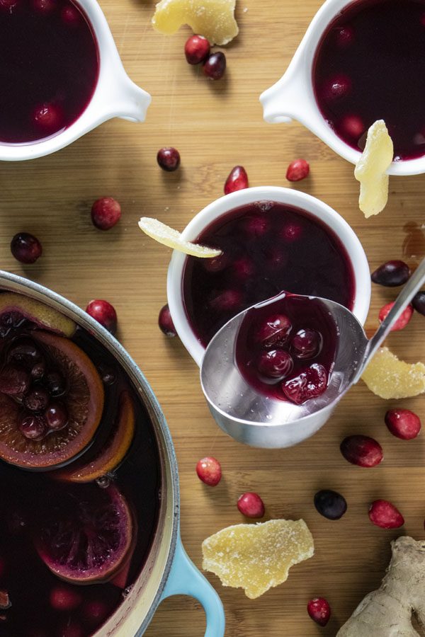 overhead photo of ladling cranberry mulled wine into a mug with cranberries and other mugs on the table