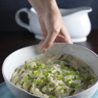 hand placing a bowl of vegan cauliflower mashed potatoes onto a table with a gravy boat in the background