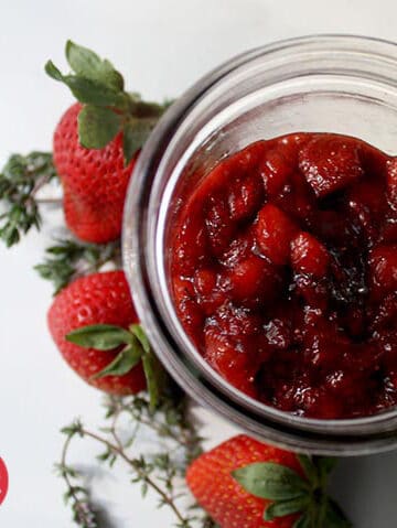jar of strawberry jam on a white table next to fresh strawberries and thyme