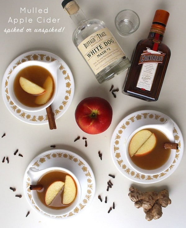 mugs of mulled apple cider on a table next to bottles of white whiskey and Cointreau, text overlay