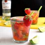 A close up of a glass of strawberry mojito garnished with a strawberry piece, lime pieces on the table