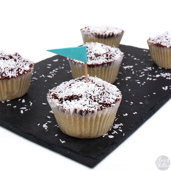 serving tray of vegan coconut cupcakes with chocolate frosting and shredded coconut on top. One cupcake has a tiny blue flag in it