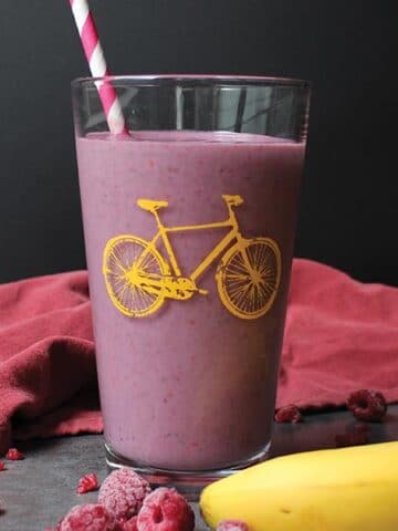 a raspberry green smoothie in a pint glass with a yellow bicycle printed on it next to frozen berries and a banana