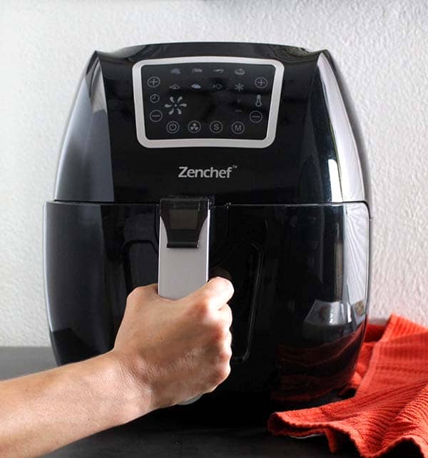 hand opening a black air fryer on a table next to a red kitchen towel