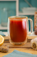 mug of tomato tea on a wooden countertop with ginger, garlic, lemon, and hot sauce around it