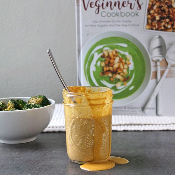Jar of easy vegan cheese sauce with cheesy broccoli and Veginner's Cookbook in the background.