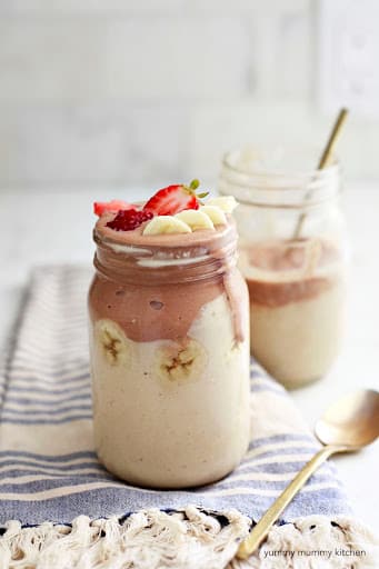 photo of a white and pink smoothie in a mason jar with strawberries and sliced banana on top. a half-drunk smoothie in the background