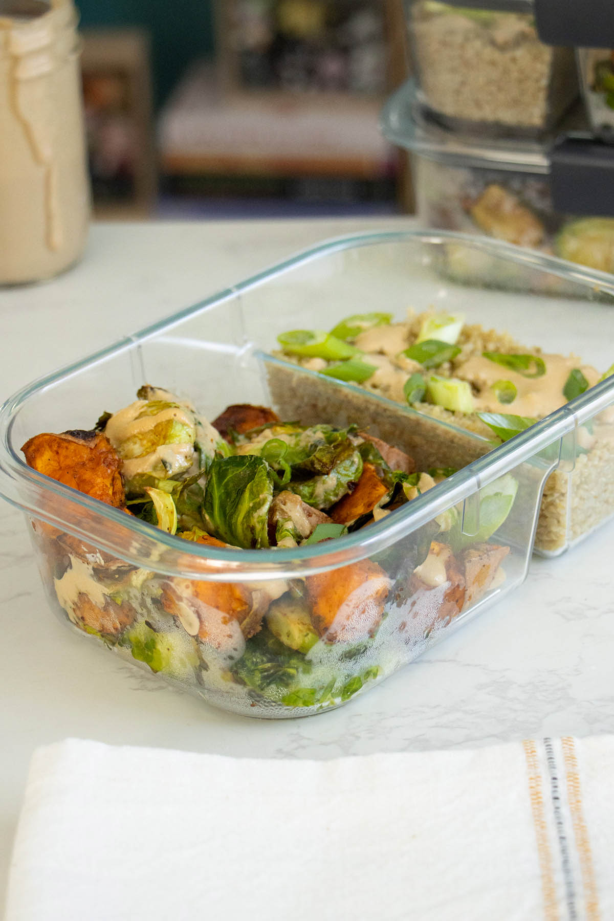 Brussels sprouts meal prep in an open lunchbox with a stack of lunchboxes behind it and the sauce in a jar on the table
