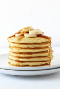 stack of vegan pancakes topped with banana slices and maple syrup on a white plate with a white background