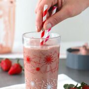 strawberry tahini smoothie in a glass with pink and red straws, strawberries and the blender in the background