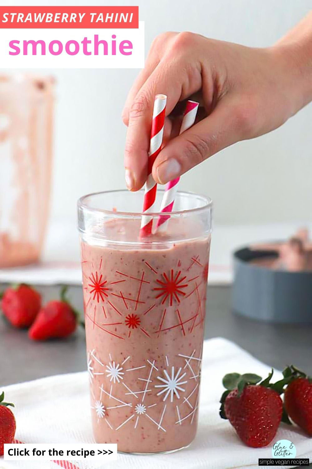 strawberry tahini smoothie in a glass with pink and red straws, strawberries and the blender in the background, text overlay