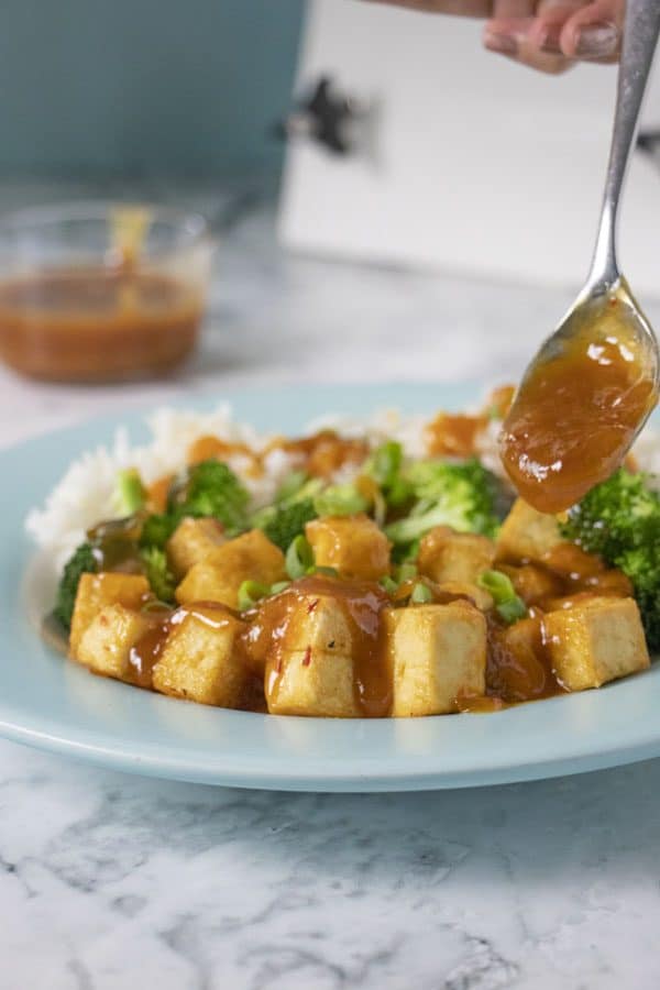 spooning sauce onto tofu, broccoli, and rice on a blue plate