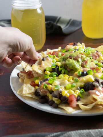 hand picking up a chip from a plate of vegan nachos with mugs of beer in the background