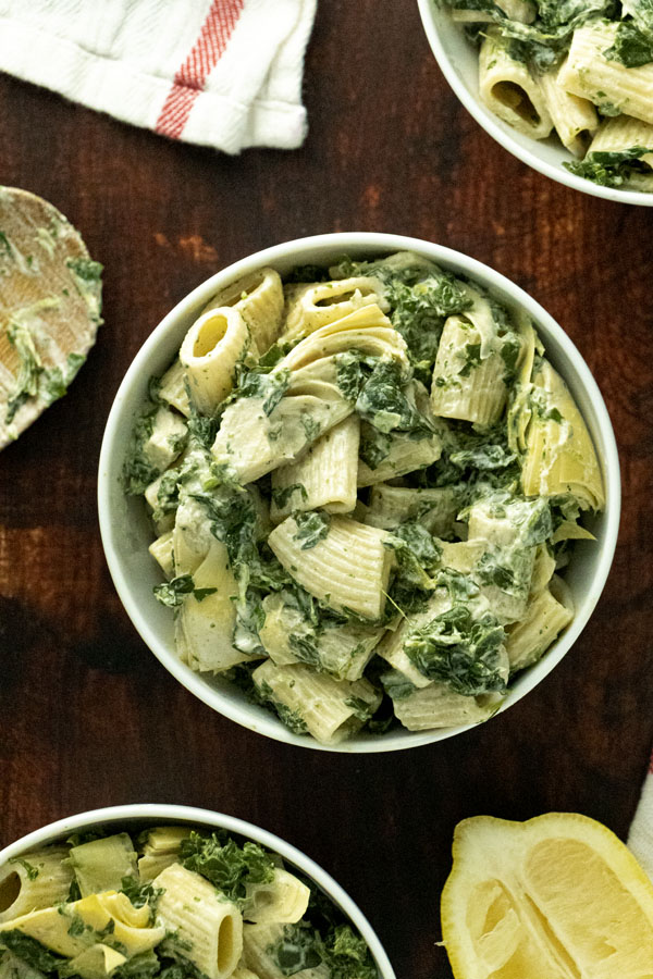 overhead photo of bowls of vegan spinach artichoke pasta on a dark wooden tabletop