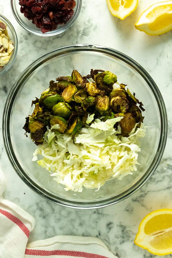 shredded cabbage and brussels sprouts in a large, glass mixing bowl with cranberries and almonds on the table