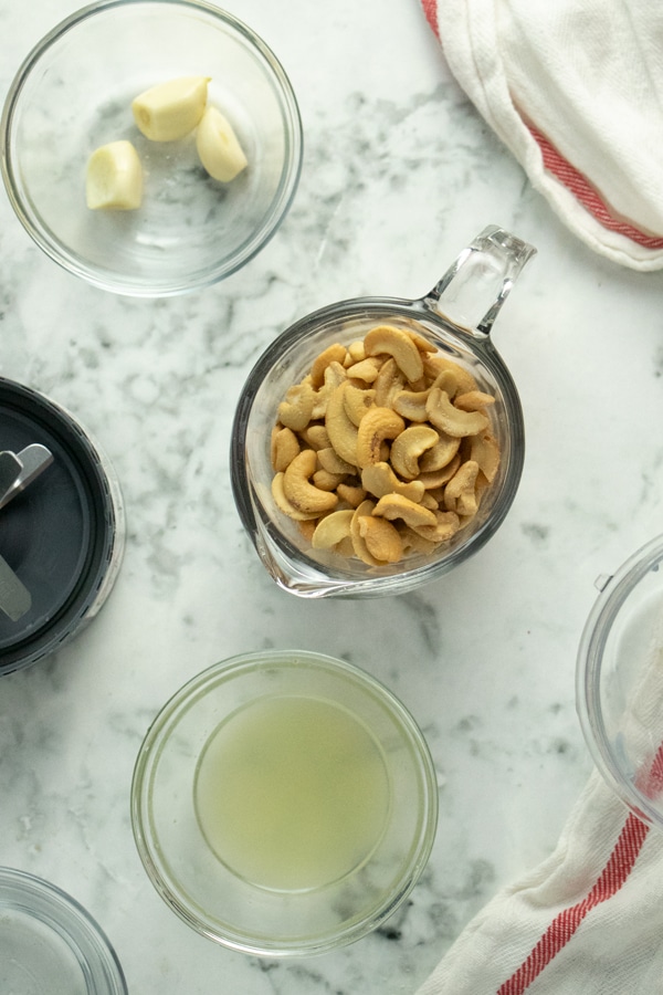 measuring cup of cashews with cups of water, lemon juice, and garlic next to it on a marble tabletop