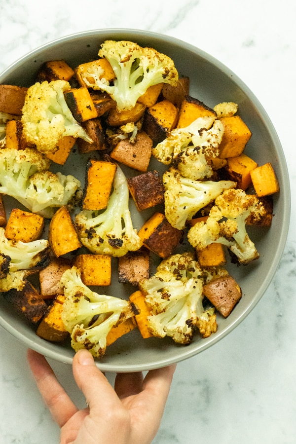 hand placing a bowl of roasted cauliflower and sweet potato onto a marble counter