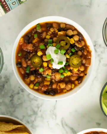 vegan bean chili topped with vegan sour cream, green onion, and jalapeno in a white bowl on a marble table
