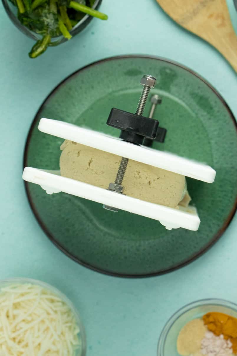 overhead photo of pressing tofu in a tofu press on a blue countertop next to bowls of frozen spinach, vegan cheese shreds, and dried spices