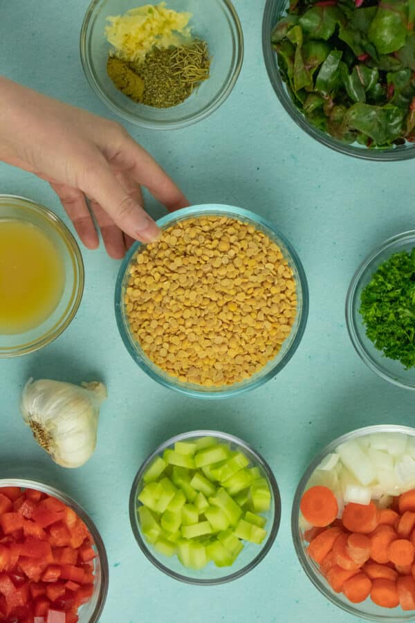 bowl of yellow split peas surrounded by bowls of chopped vegetables and herbs on a blue table