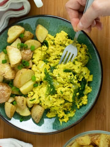 A plate of tofu scramble with a side of breakfast potatoes sitting on top of a wooden table