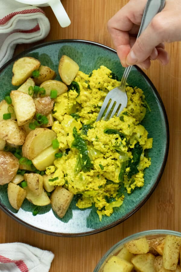 A plate of tofu scramble with a side of breakfast potatoes sitting on top of a wooden table