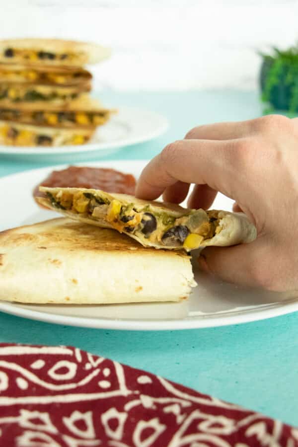 hand placing a vegan quesadilla with a bite out of it onto a white plate. Stack of more quesadillas in the background
