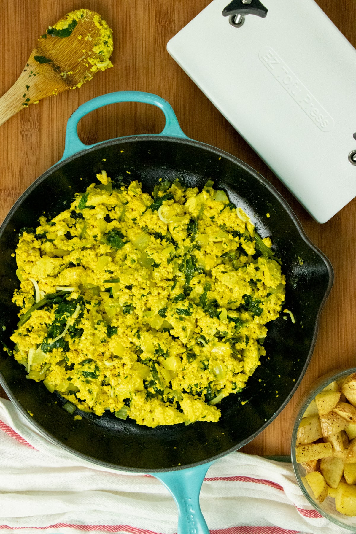 overhead photo of a basic tofu scramble with spinach in a skillet next to a tofu press, a serving spoon, and a bowl of crispy potatoes