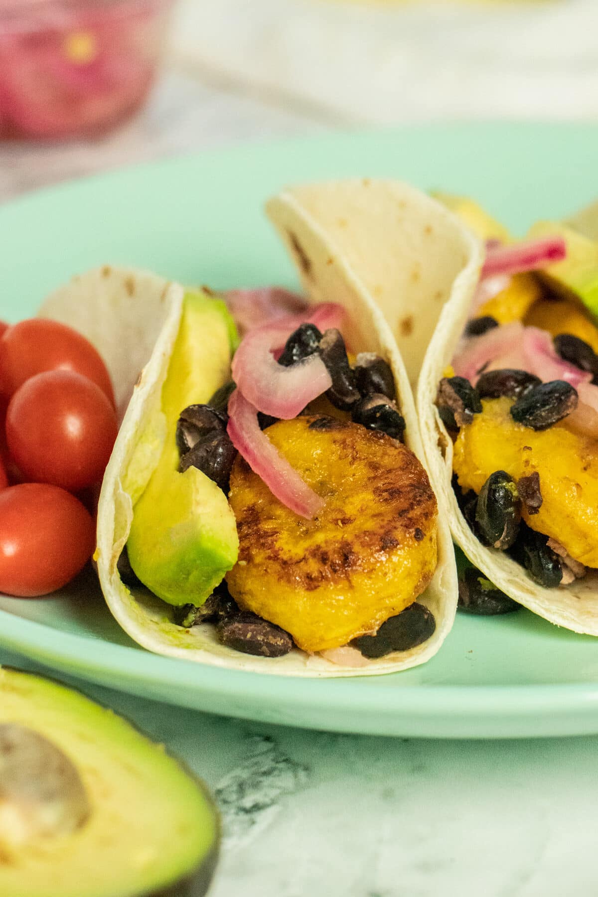 close-up of plantain tacos with red onion, black beans, and sliced avocado. Grape tomatoes on the side