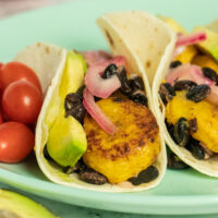 close-up of plantain tacos with red onion, black beans, and sliced avocado. Grape tomatoes on the side