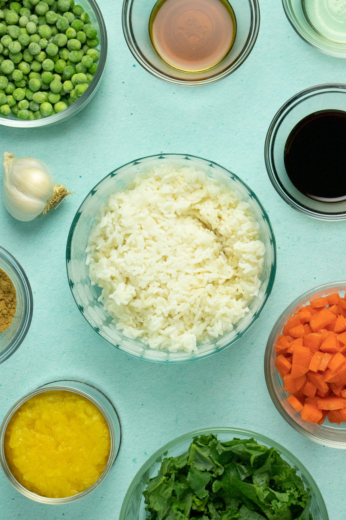 overhead photo of a blue table with glass bowls of rice, vegetables, and spices and a can of pineapplee