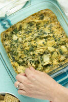 photo of vegan spinach artichoke dip in the baking pan. A hand is dipping a cracker into the pan.