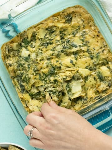 photo of vegan spinach artichoke dip in the baking pan. A hand is dipping a cracker into the pan.