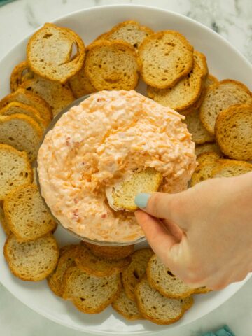 hand dipping a cracker into a serving bowl of vegan pimento cheese