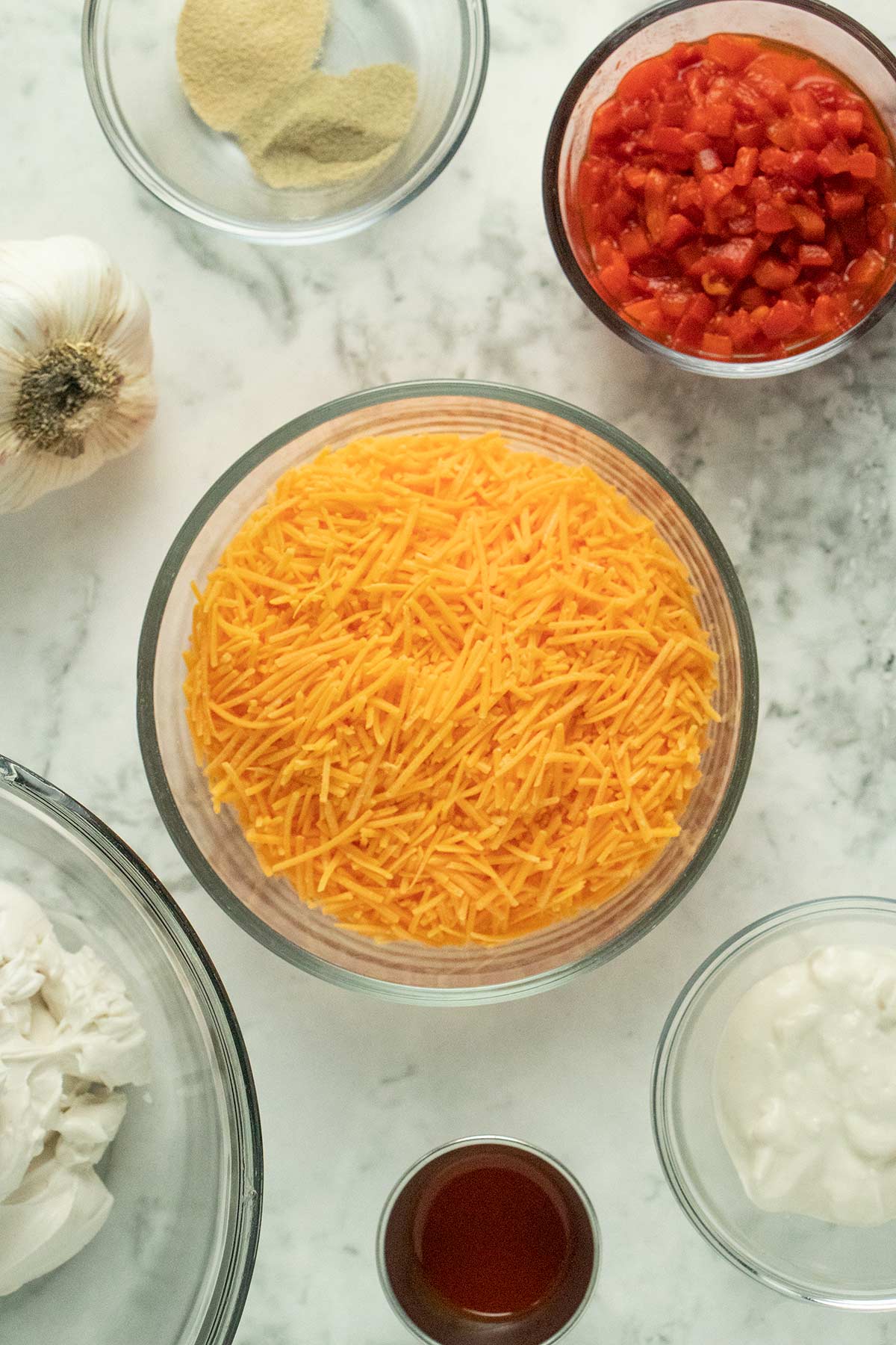 bowl of vegan cheddar shreds with other bowls of ingredients around it on a marble countertop