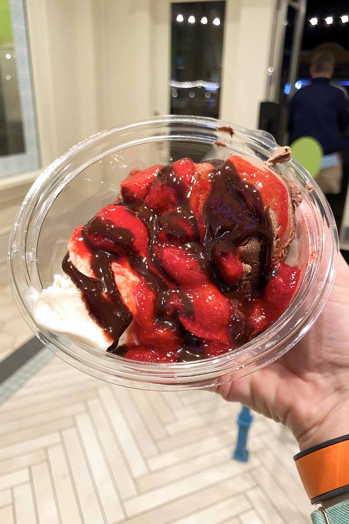 hand holding a sundae with chocolate sauce, strawberry syrup, and strawberry pieces on it