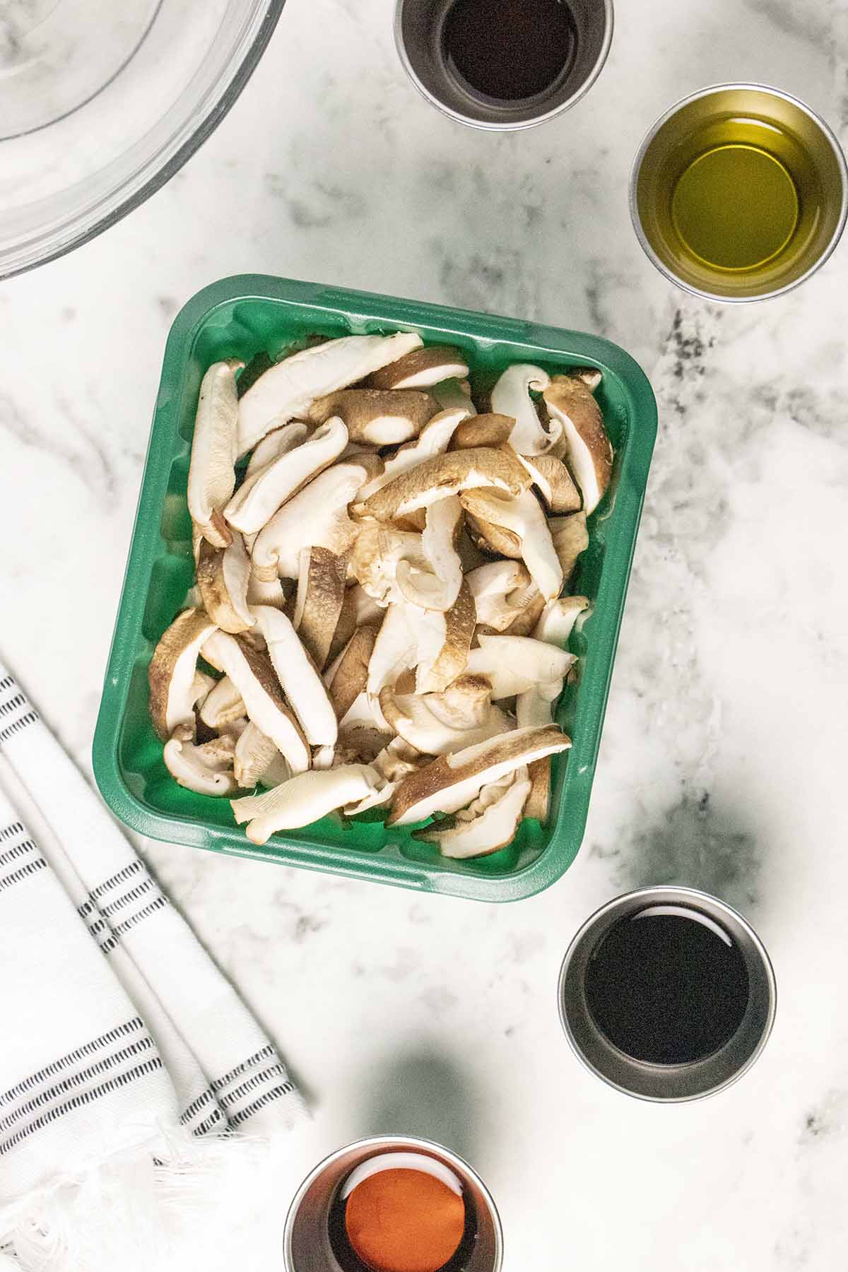 shiitake mushrooms in a green container on a marble table surrounded by cups of liquid ingredients
