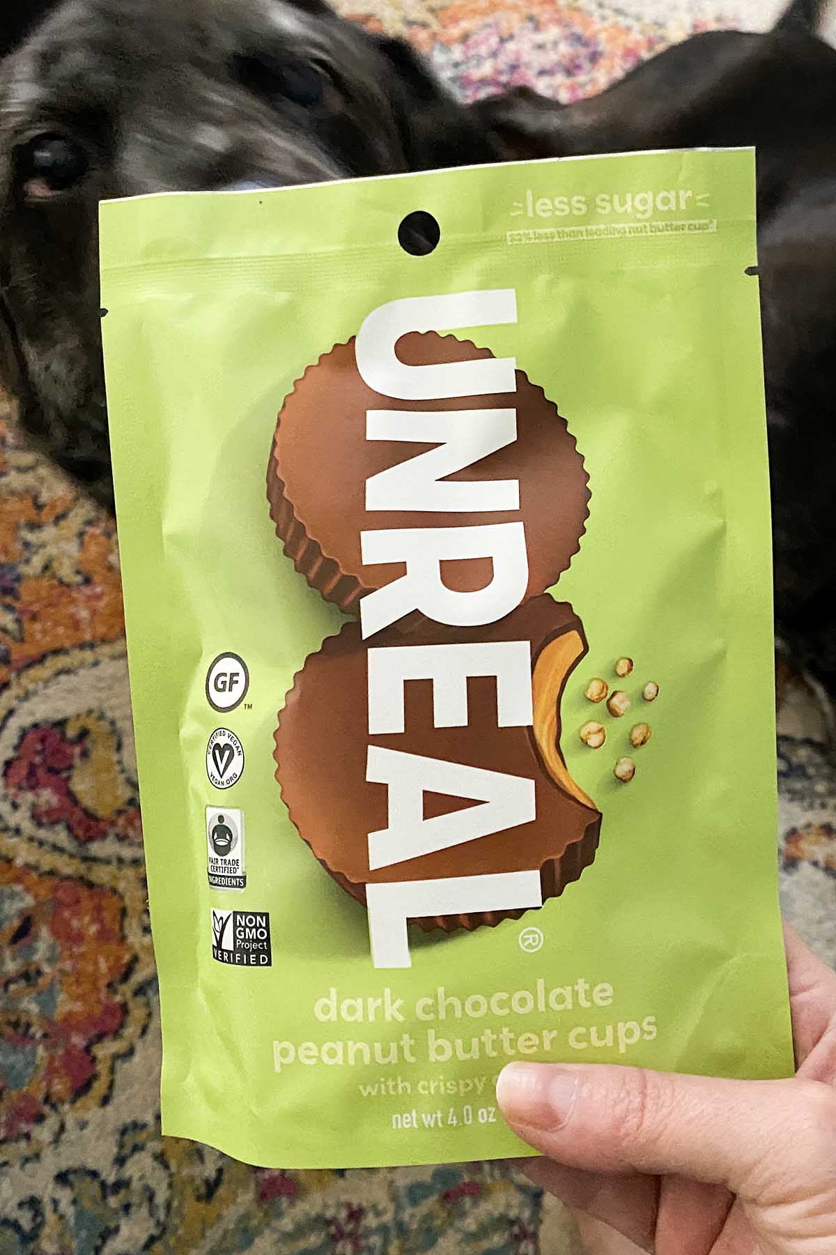 hand holding a bag of Unreal Peanut Butter Cups, a dog is photo bombing this shot