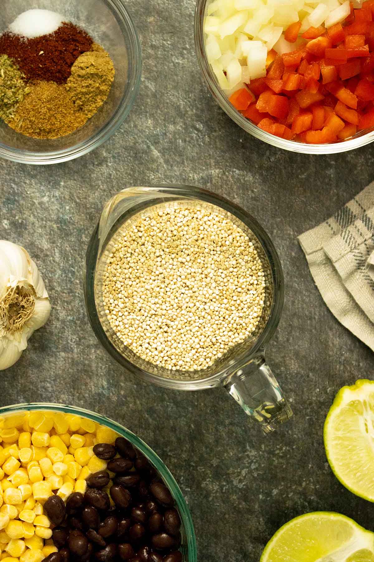 cup of quinoa surrounded by bowls of beans and corn, spices, and veggies on a gray table