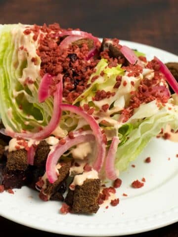 vegan wedge salad with dark croutons, pickled onions, bacon, and creamy dressing on a white plate
