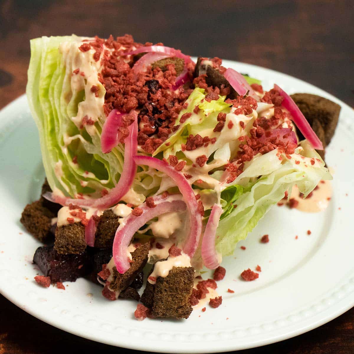 vegan wedge salad with dark croutons, pickled onions, bacon, and creamy dressing on a white plate