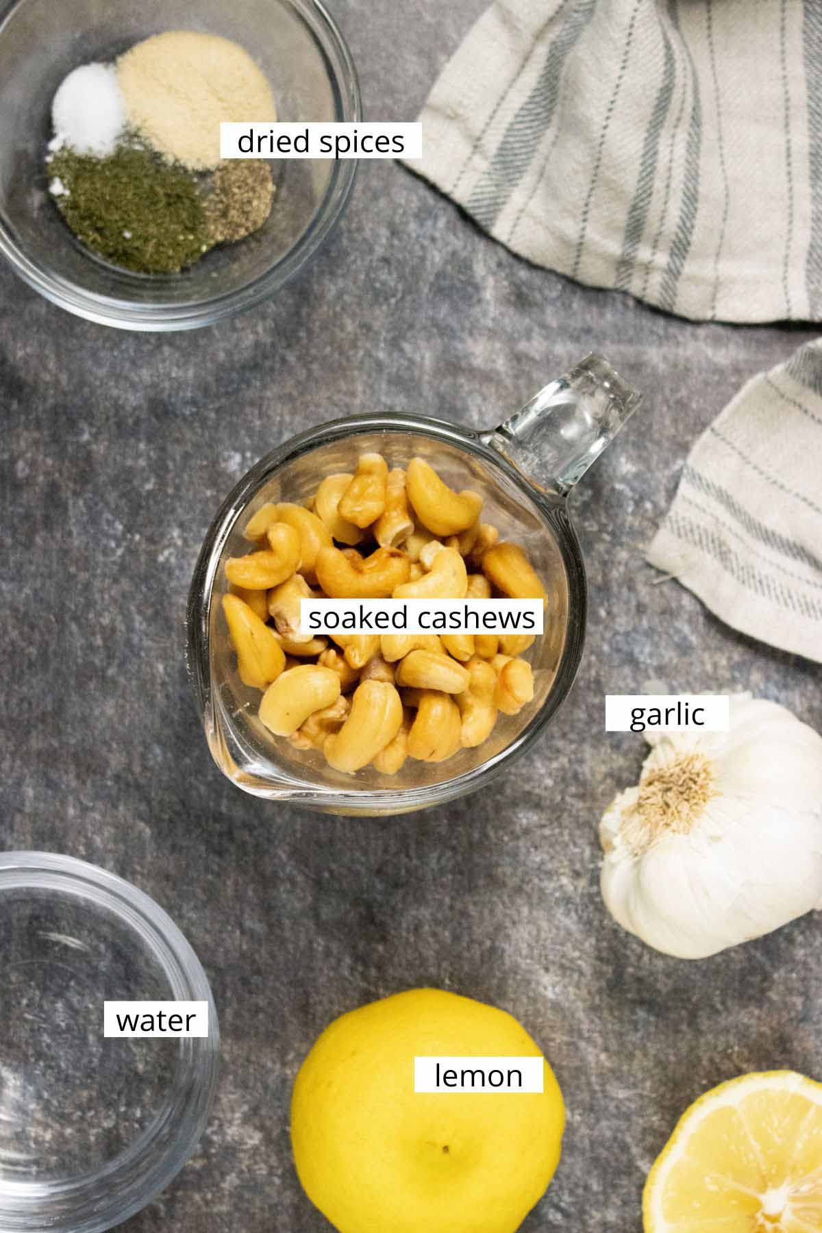 cashews, garlic, lemon, water, and spices in bowls on a slate table with text labels on each ingredient