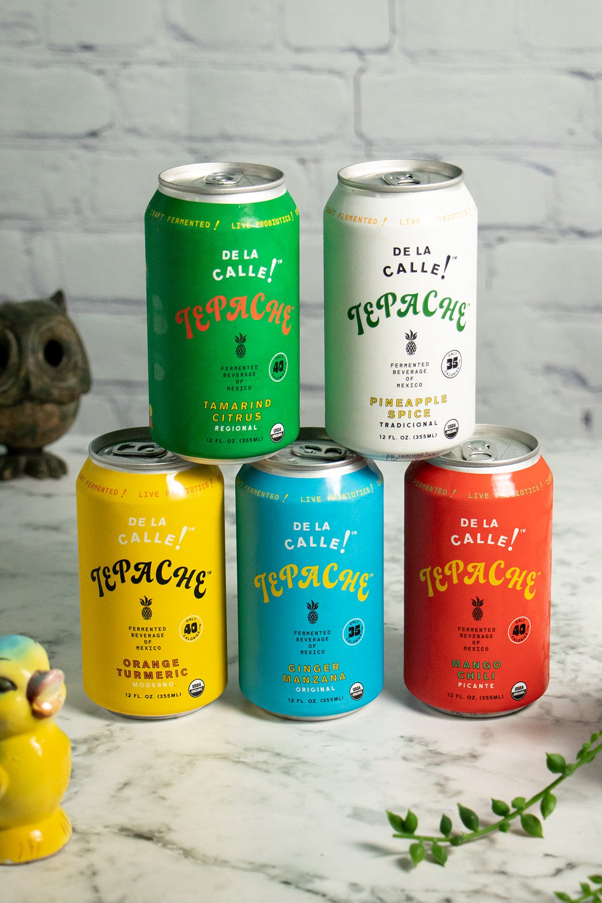 colorful cans of De La Calle Tepache stacked up on a marble table surrounded by cute tchotchkes