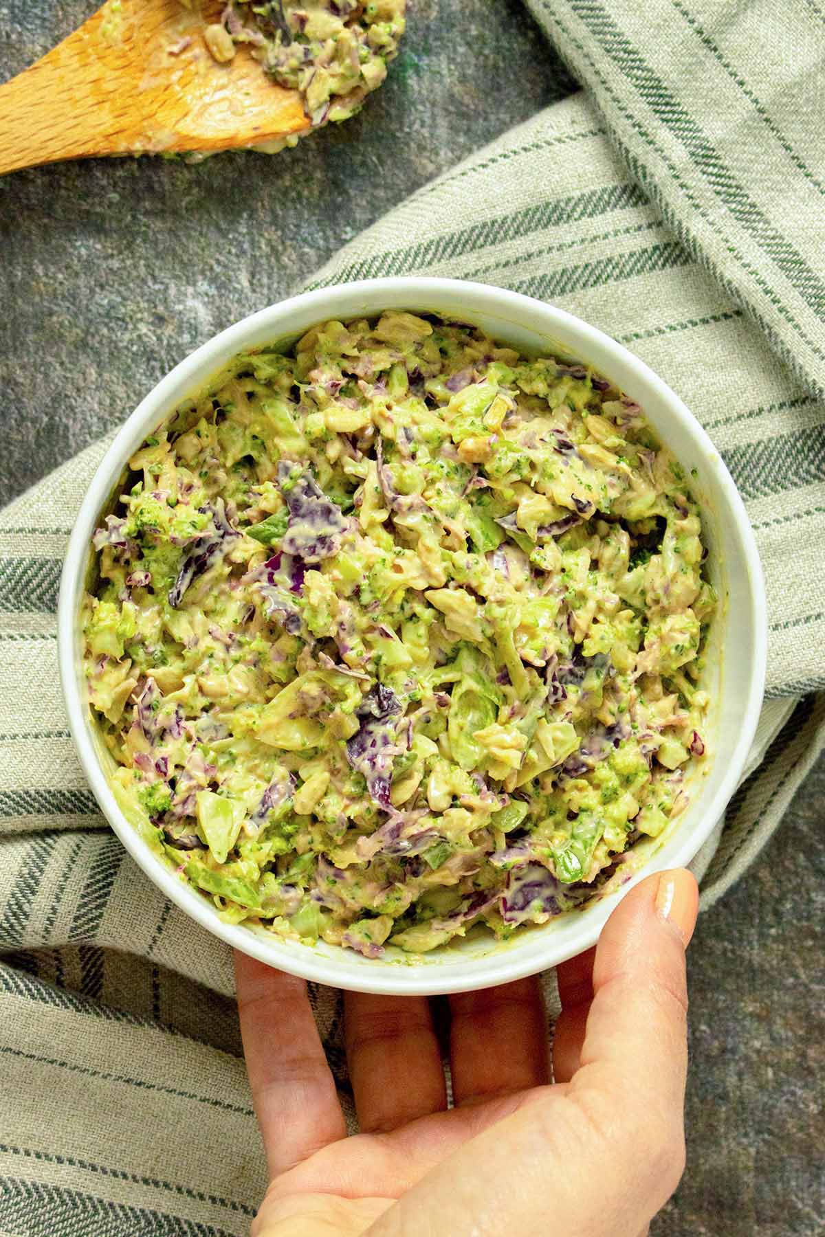 hand placing a bowl of broccoli slaw on a slate table next to a serving spoon