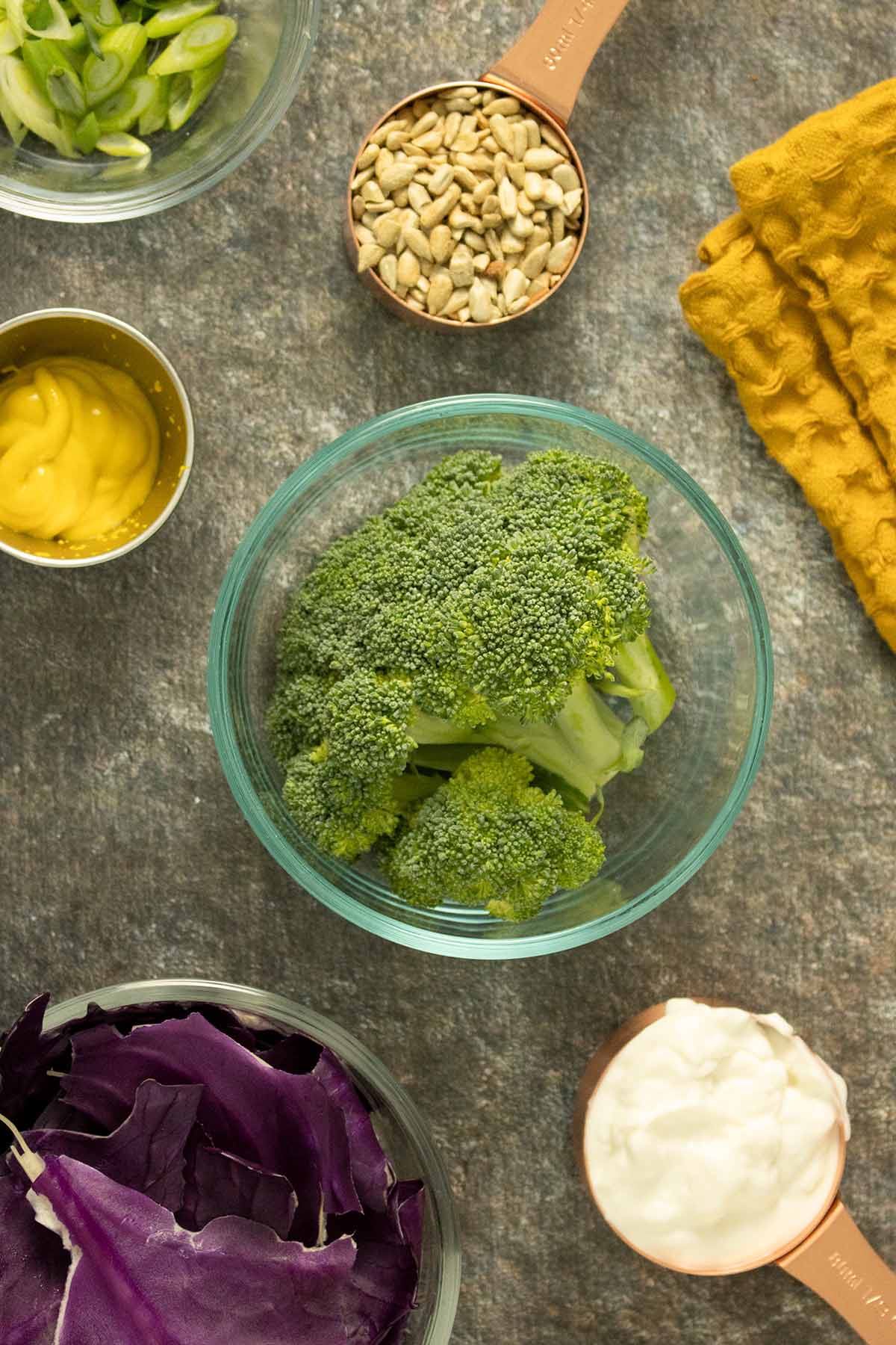 broccoli, cabbage, and other slaw ingredients in cups and bowls on a slate table