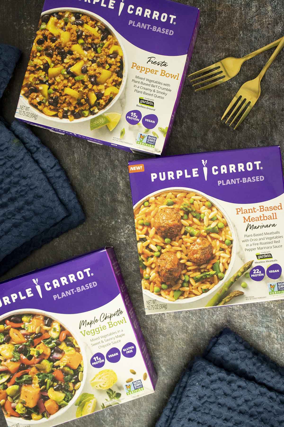 3 boxes of Purple Carrot Frozen Meals on a slate table with cloth napkins and gold forks