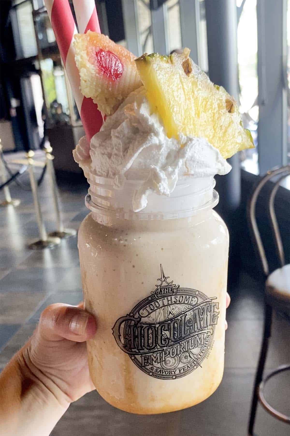 hand holding a vegan milkshake in a mason jar with whipped cream, pineapple, and a whole slice of cake on it