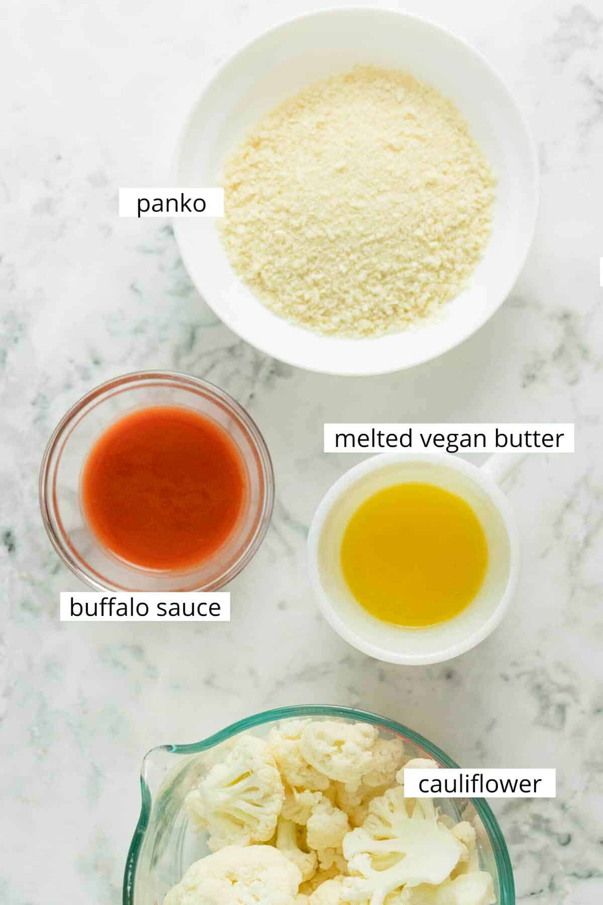 panko, buffalo sauce, vegan butter, and cauliflower florets in bowls on a marble table. All labeled with text