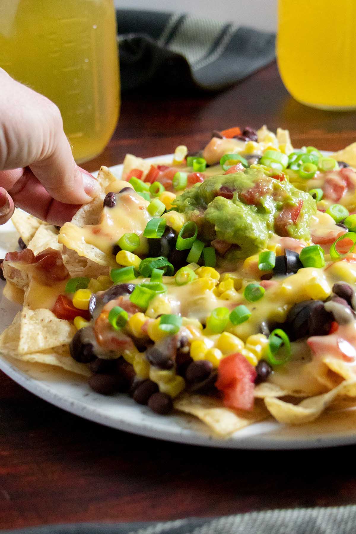 hand grabbing a chip from a plate of cheesy vegan nachos, beers in the background