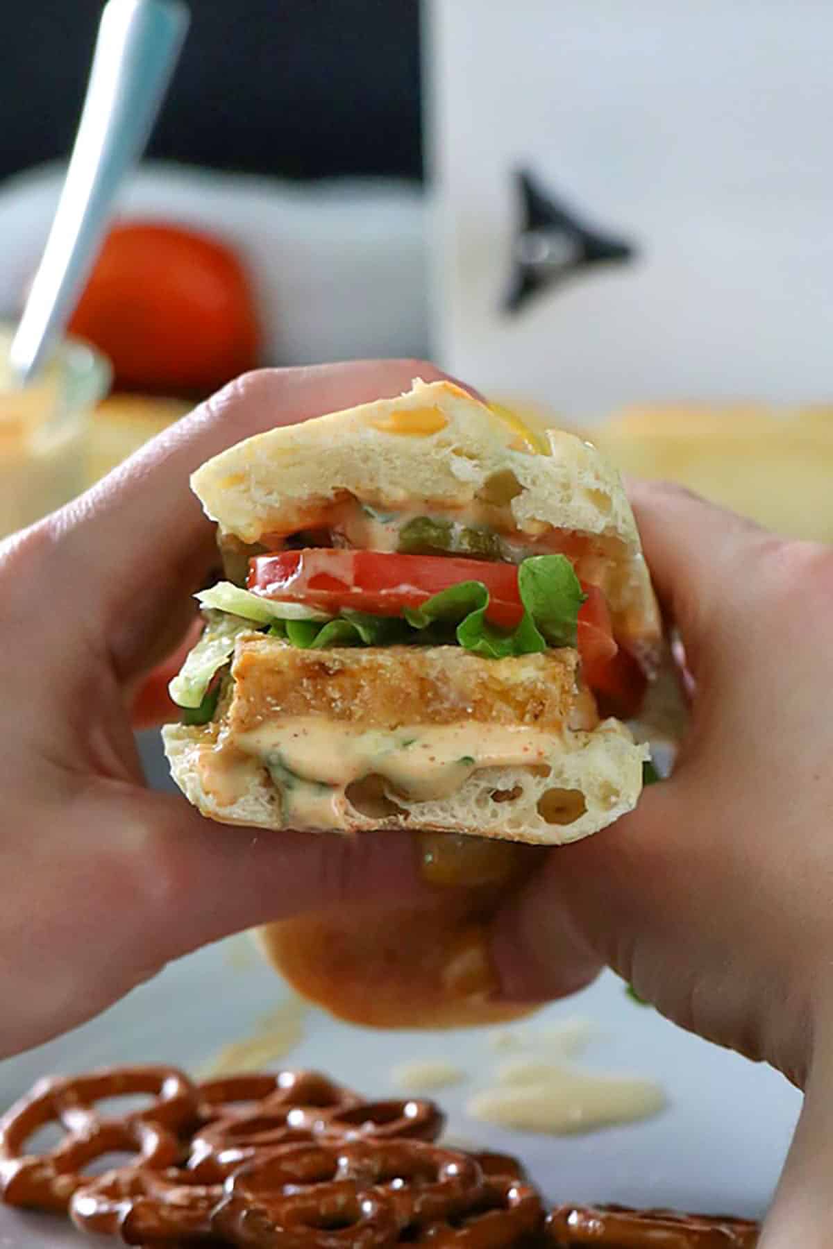 A close up of hands holding baked vegan po' boy with lettuce, tomato, and pickles